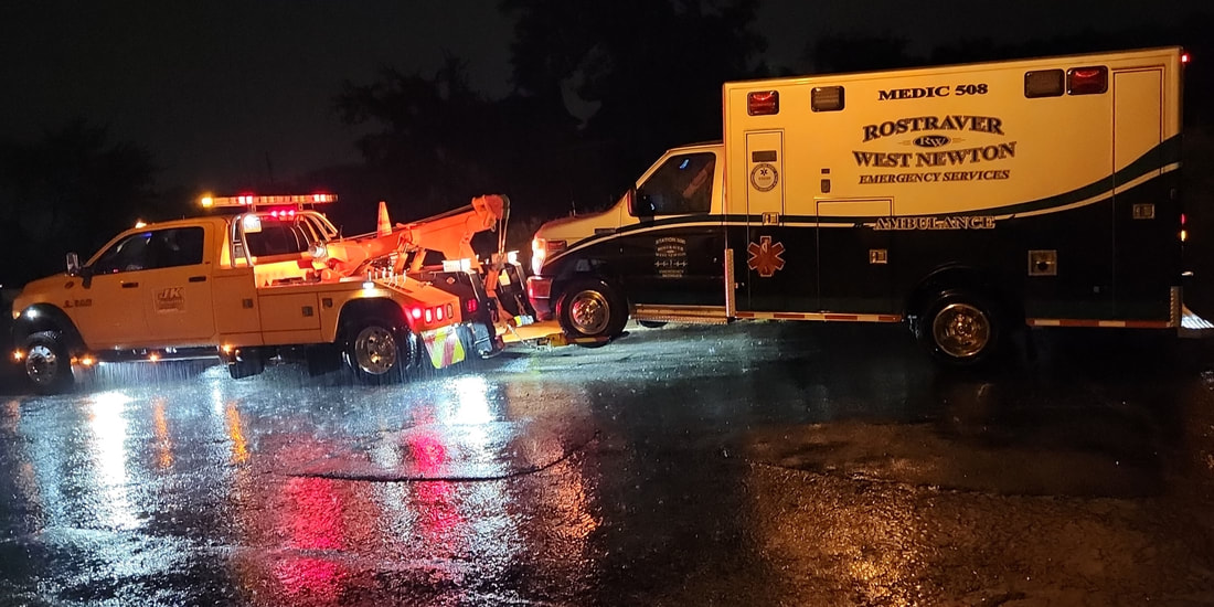 working at night during a rainstorm, JK Towing's tow truck uses its wheel-lift boom to tow an ambulance by its front wheels